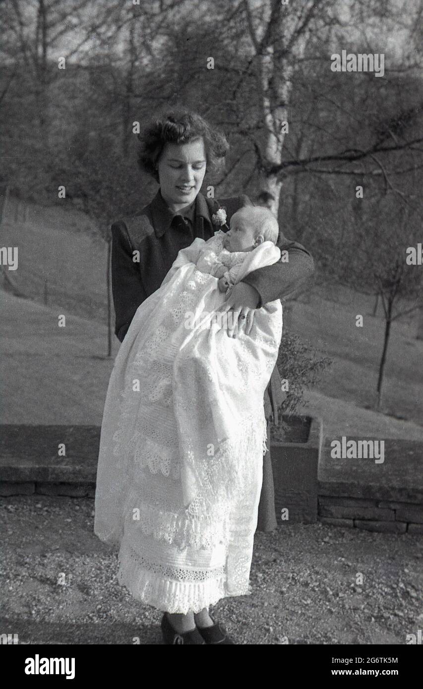 Anni '50, storica, una nuova madre in piedi fuori tenendo il suo bambino  avvolto in un lungo cotone o pizzo modellato scialle, in occasione speciale  del battesimo del bambino, Inghilterra, Regno Unito.