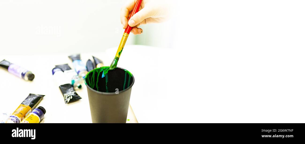 la mano dell'artista lava un pennello con vernici in un bicchiere d'acqua, uno strumento dell'artista, la creatività, il disegno, la formazione al disegno, scuola d'arte, banner Foto Stock