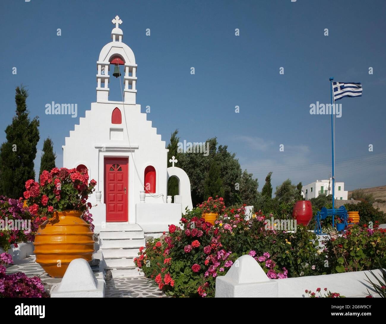 Piccola cappella greca con campanile e bandiera greca circondata da giardino fiorito in campagna con cielo blu sull'isola di Mykonos in primavera, Grecia Foto Stock