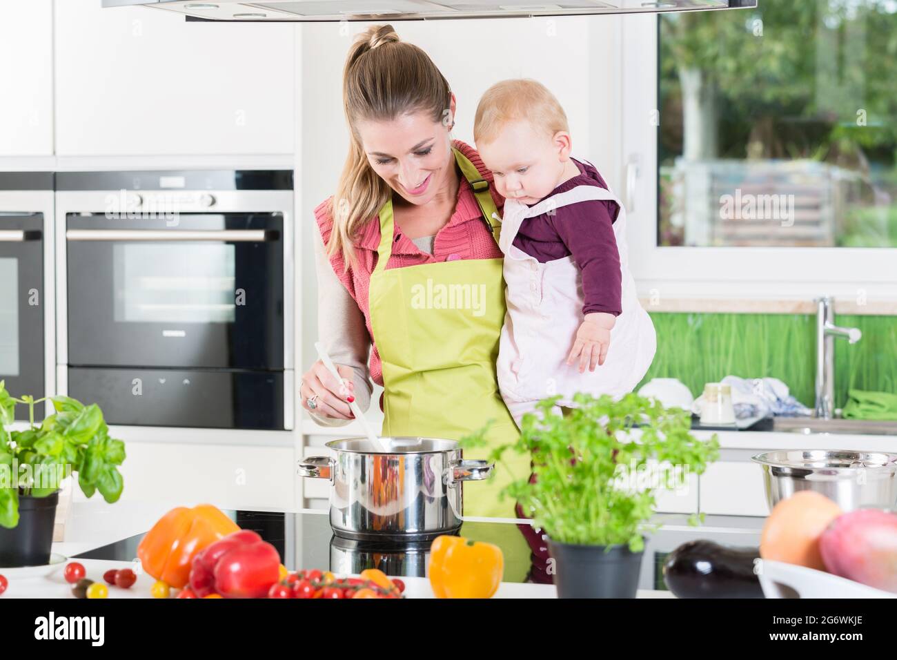 Mamma cucinando con il bambino nel braccio nella sua cucina domestica Foto Stock