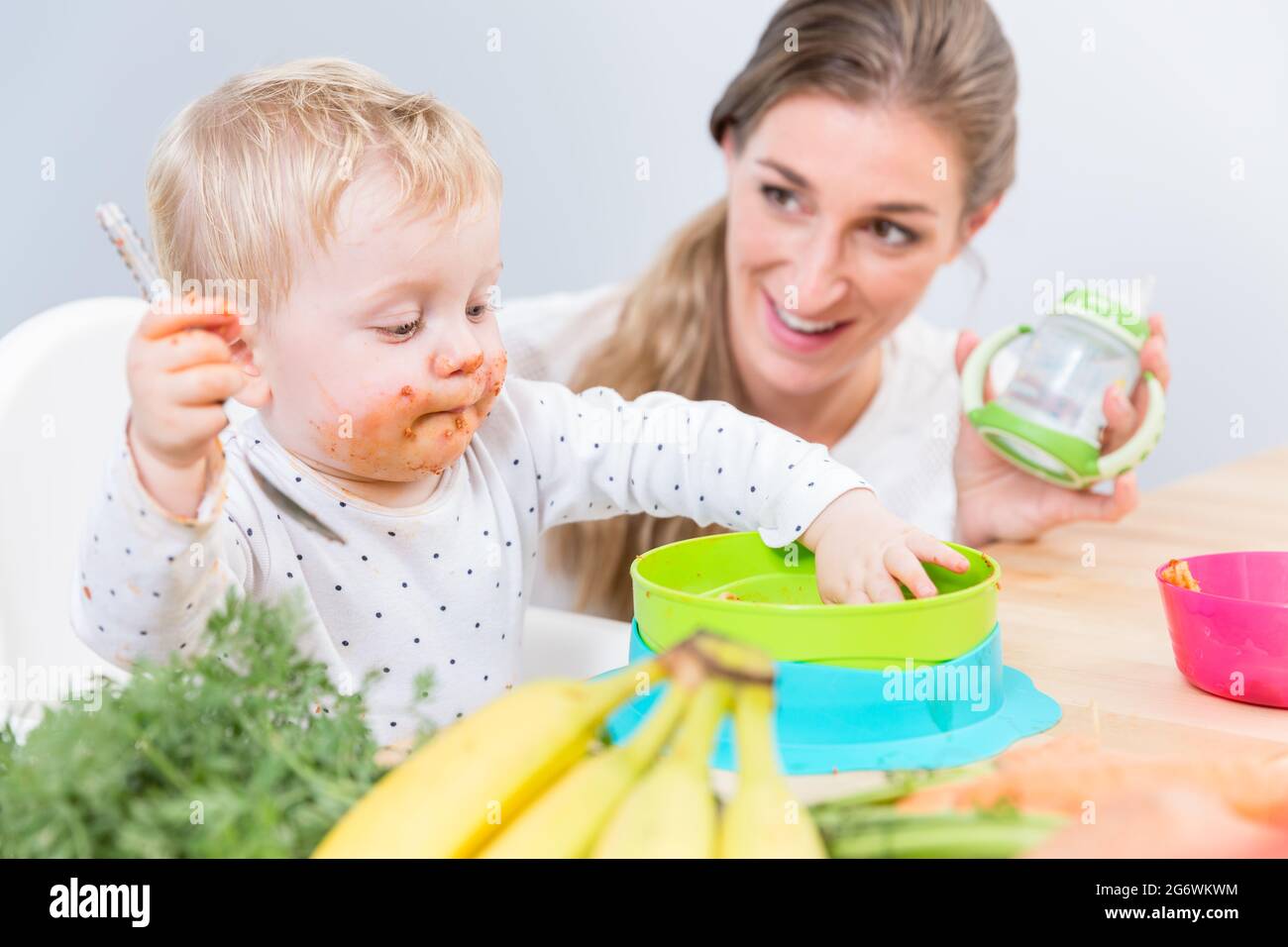 Ritratto di una bambina carina e divertente seduta su una sedia alta, mentre mangia con le mani cibo solido da una ciotola di plastica accanto alla sua madre dedicata Foto Stock