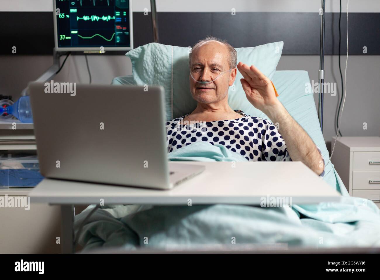 Ritratto dell'uomo anziano che saluta la famiglia a una macchina fotografica del portatile che si trova nel letto dell'ospedale, dopo la diagnosi della malattia, che respira con il tubo dell'ossigeno. Moderno eqipment per il monitoraggio della frequenza cardiaca paciente durante il recupero. Foto Stock