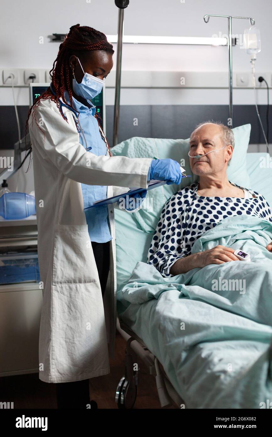 Medico afroamericano con maschera chirurgica che legge la diagnosi dalla clipboard a malato malato malato paziente anziano che si posa a letto, respirando con l'aiuto della maschera di ossigeno, ascoltando discutere con il personale medico circa il recupero. Foto Stock