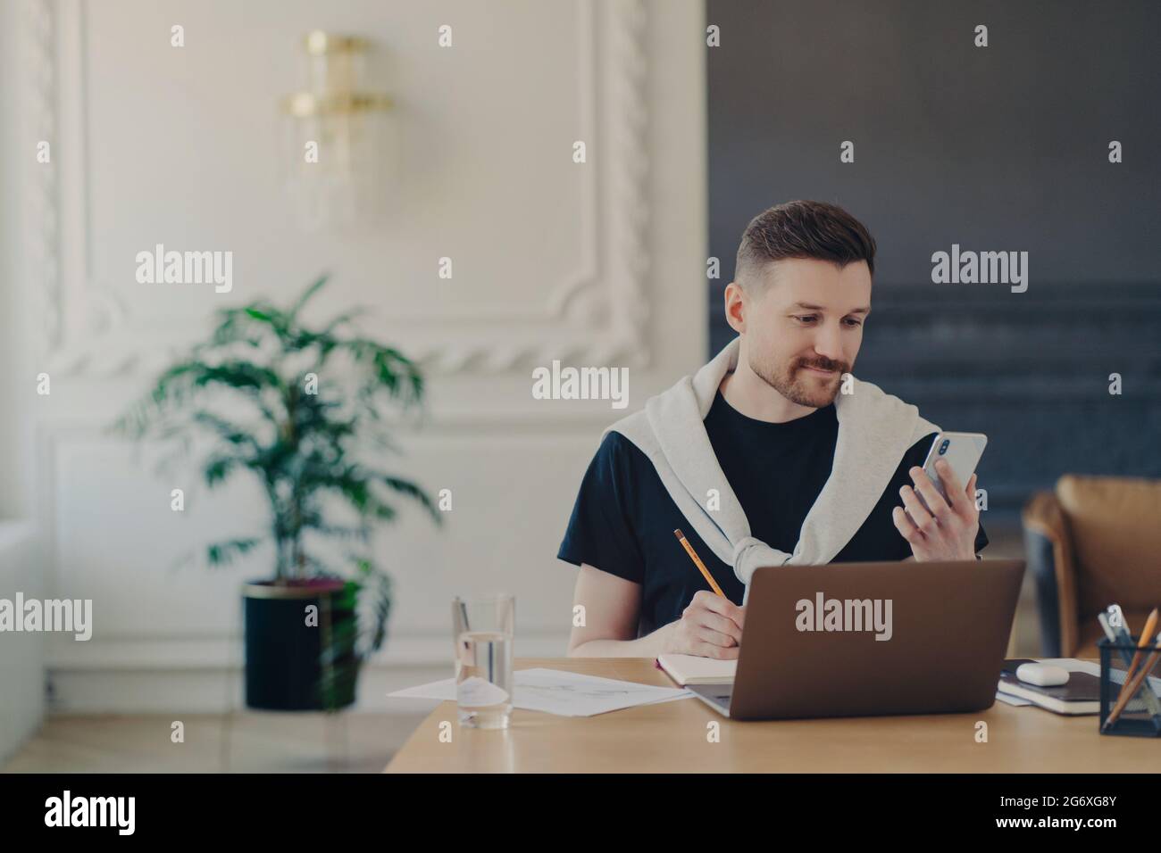 Imprenditore maschile che studia online, utilizzando lo smartphone mentre lavora sul suo posto di lavoro in un ufficio moderno Foto Stock