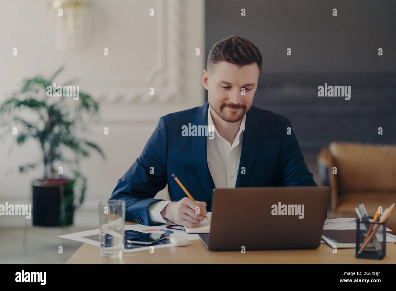 Il colpo interno di lavoro maschile bello riscrive le informazioni dal notepad nel diario siede alla scrivania vestita in abiti formali spende il tempo per la ricerca Foto Stock