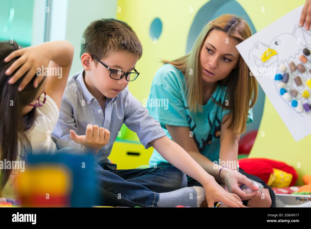 Carino pre-scuola ragazzo indossa occhiali imparando a leggere da un libro per bambini aiutati da un apposito asilo assistente insegnante Foto Stock