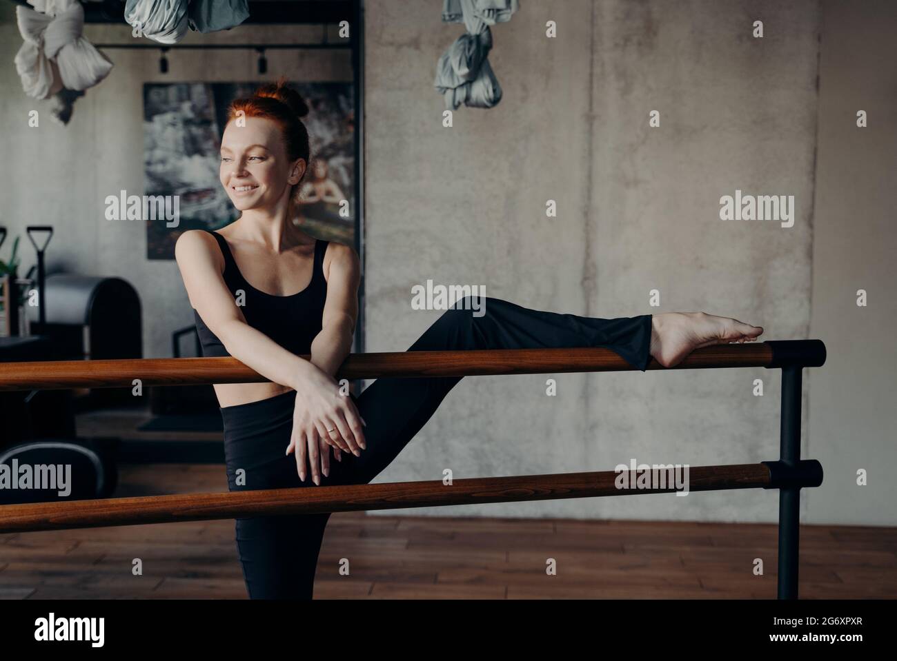 Snella donna dai capelli rossi sorridente che allunga la gamba sul balletto barre in classe Foto Stock