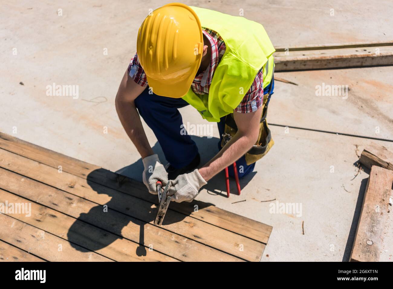 Vista ad alto angolo di un operaio con collare blu che indossa elmetto giallo, giubbotto di sicurezza e guanti mentre si utilizza un martello durante il lavoro sul cantiere Foto Stock