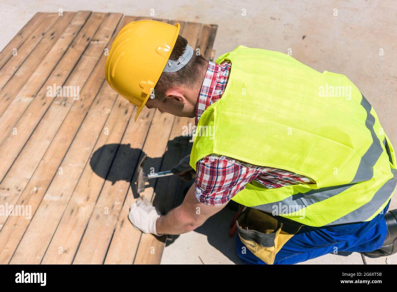 Vista ad alto angolo di un operaio con collare blu che indossa elmetto giallo, giubbotto di sicurezza e guanti mentre si utilizza un martello durante il lavoro sul cantiere Foto Stock