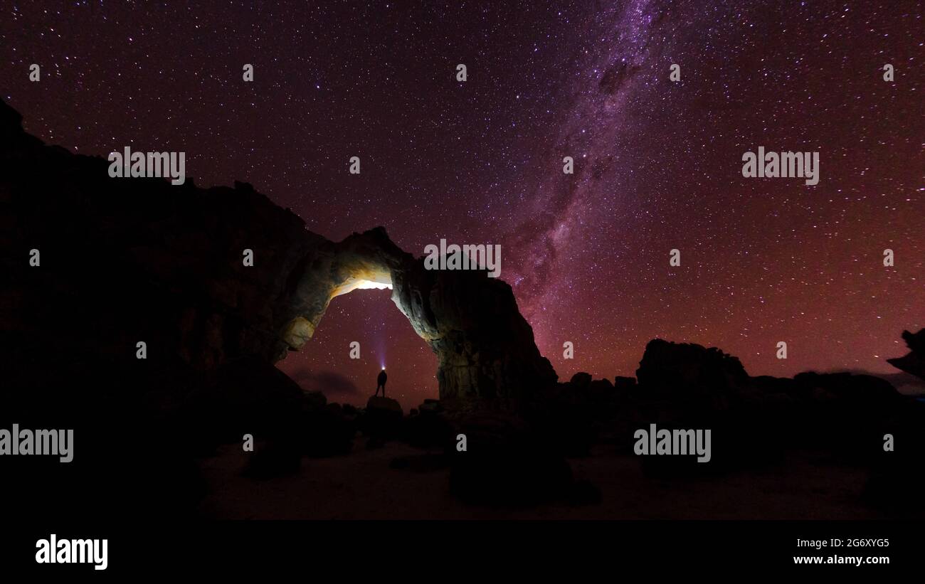 Una persona si trova sotto un arco di roccia, fissando il cielo notturno pieno di stelle. Foto Stock