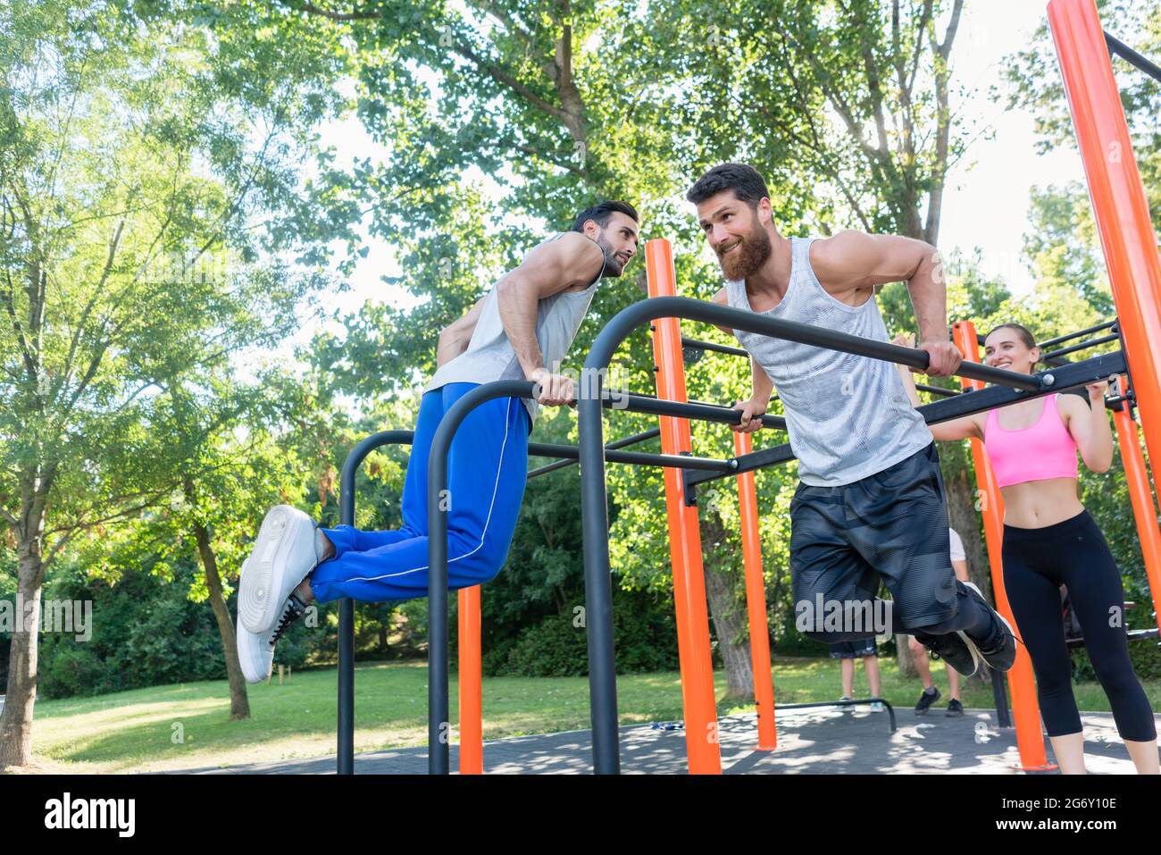 Due giovani uomini forti e motivati che fanno un'attività fisica per la parte superiore del corpo, durante l'allenamento impegnativo con il loro amico in un parco fitness all'aperto WIT Foto Stock
