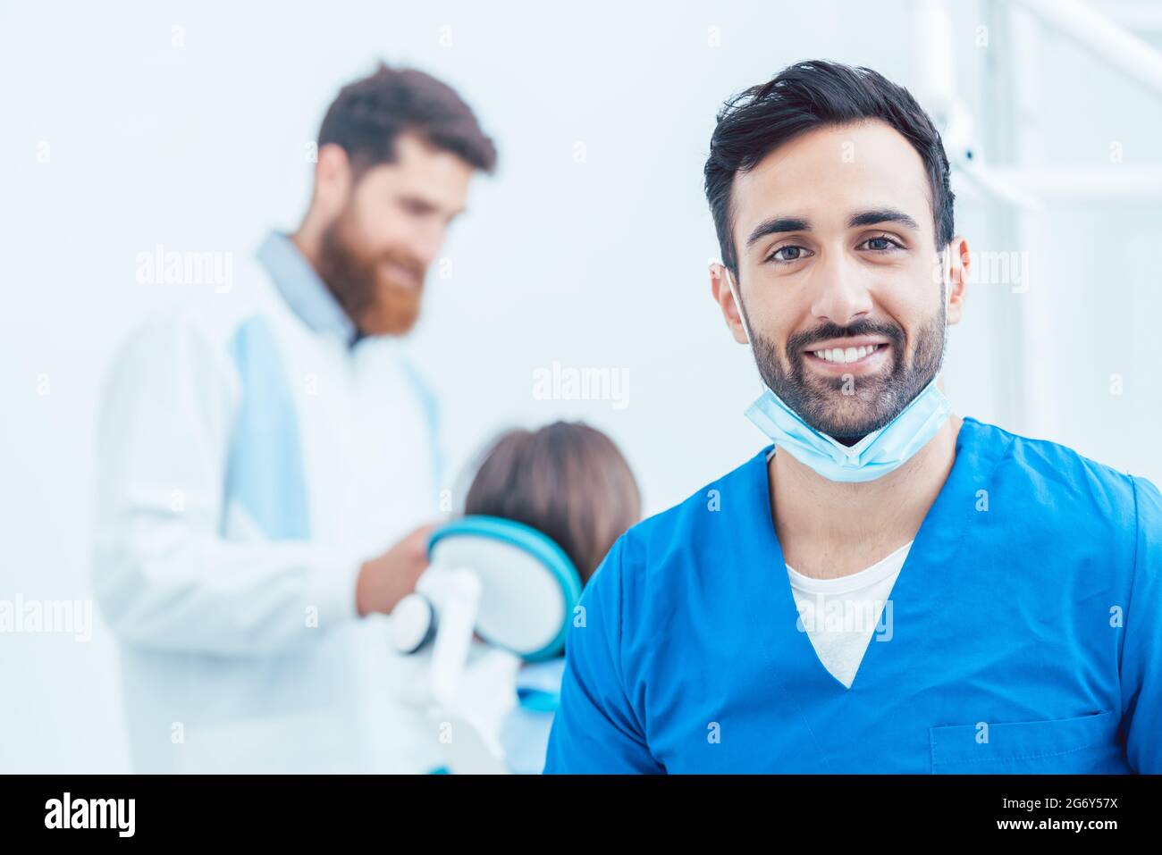 Ritratto di un sicuro chirurgo dentale indossando uniforme protettiva blu e maschera chirurgica mentre si guarda la fotocamera in un moderno studio dentistico con reliab Foto Stock