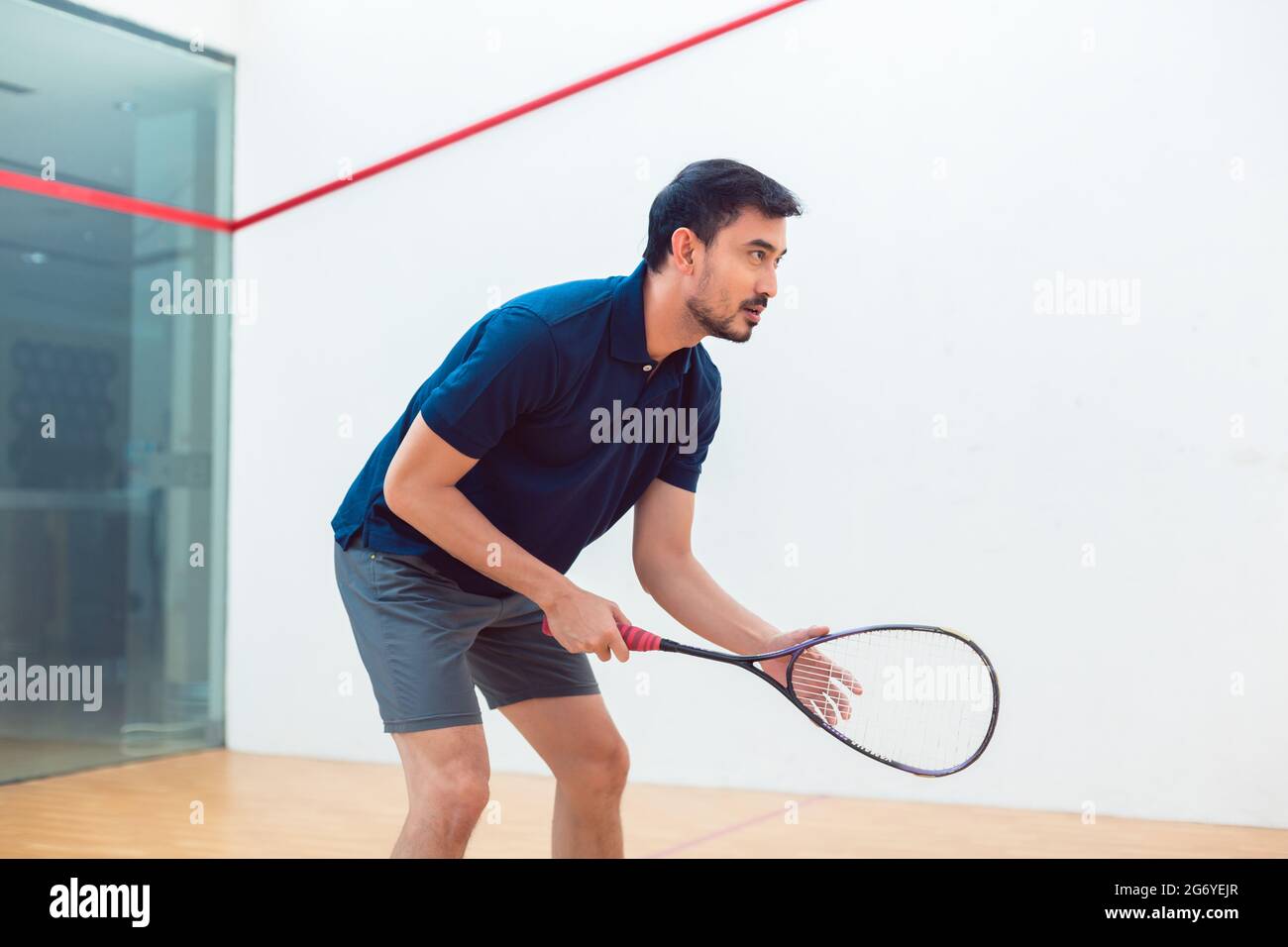 Vista laterale di un giovane giocatore di squash che tiene la racchetta mentre si è in piedi con le ginocchia piegate durante la partita in interni su un campo professionale Foto Stock