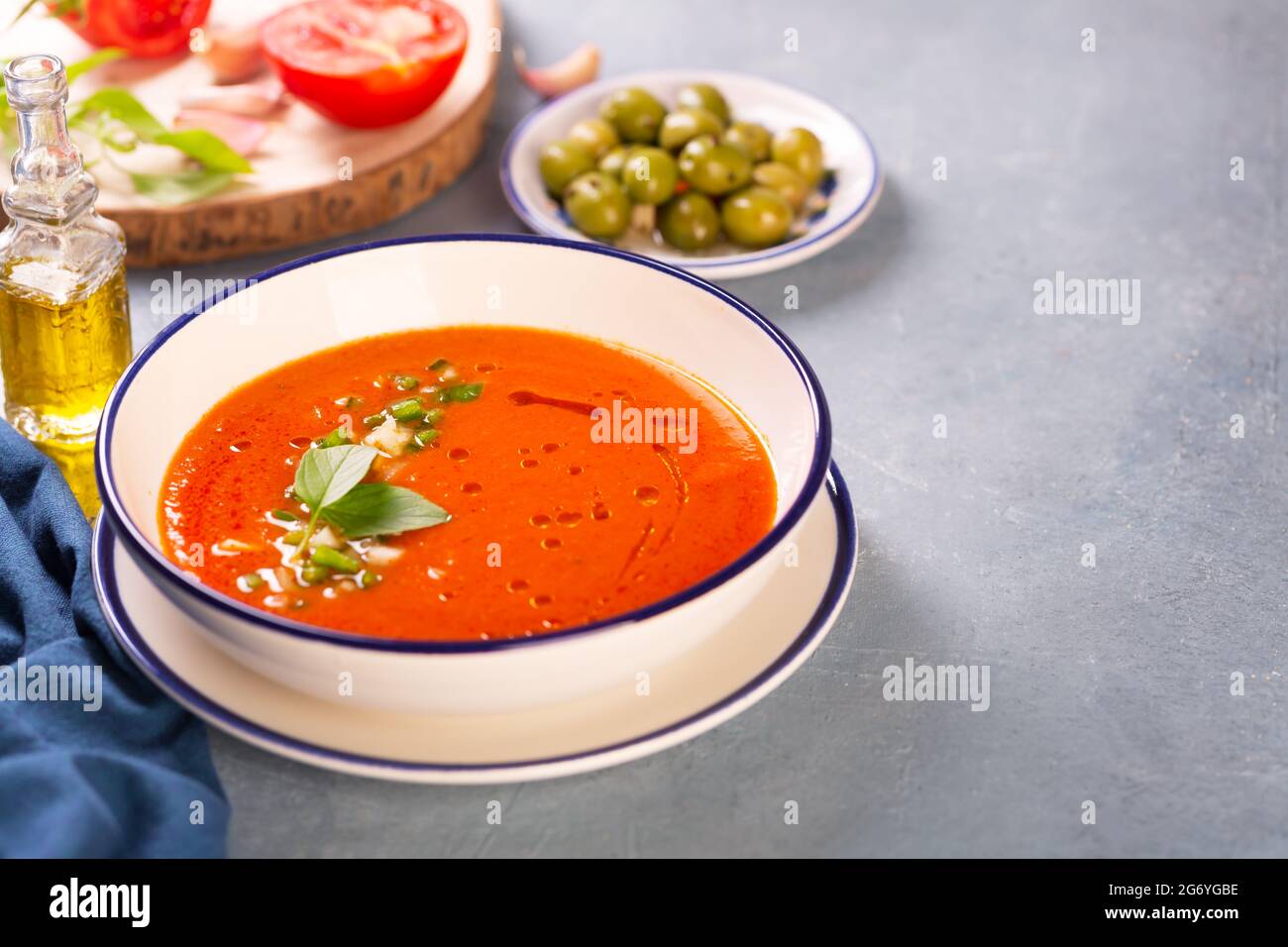 Un piatto di gazpacho spagnolo tradizionale sullo sfondo di ingredienti come pomodori, olive, olio d'oliva, aglio e spazio copia di stoffa blu Foto Stock