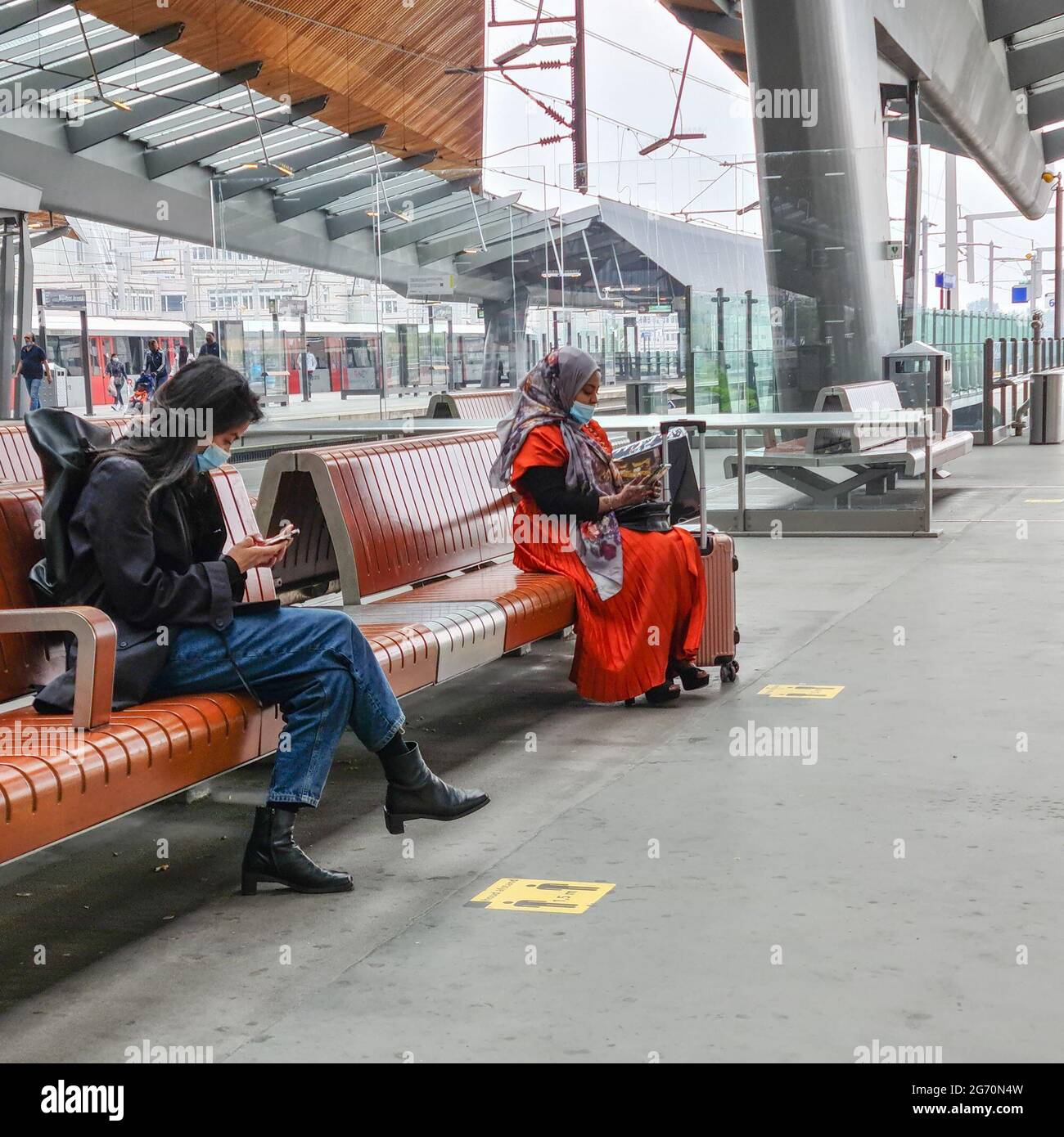 Amsterdam, Paesi Bassi, 25 giugno 2021 : due Signore di diverse culture, indossando maschere e controllando i loro telefoni seduti sulle panchine nella stazione di Amsterdam Bijlmer Arena, in attesa del loro treno. I contrassegni gialli sul terreno indicano una distanza di 1,5 metri, una delle regole di base durante la pandemia di covidio. Foto Stock
