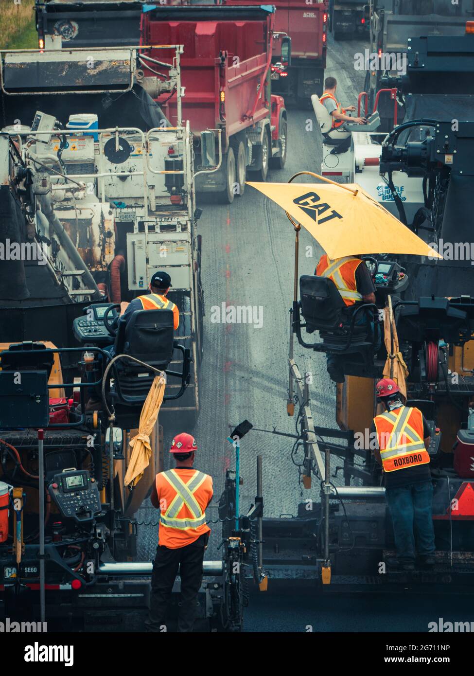 Uomini che lavorano più persone con cappelli duri e giubbotti arancioni riflettenti che utilizzano macchine per pavimentazione in asfalto CAT e dumper sulla strada statale 25 in Quebec. Foto Stock