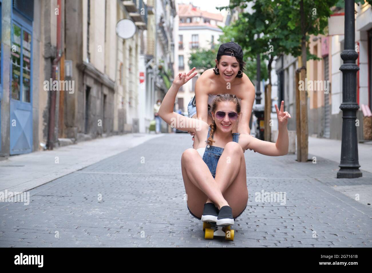 Una donna che spinge la sua amica da dietro su uno skateboard. Due amici di sesso femminile che giocano in strada. Foto Stock