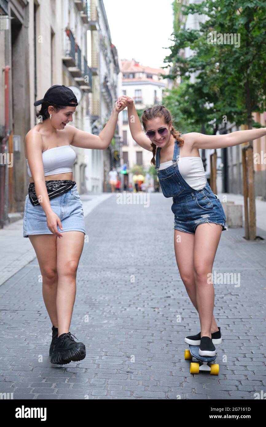 Una donna che aiuta la sua amica a stare in piedi su uno skateboard. Due amiche che si divertono in strada. Foto Stock