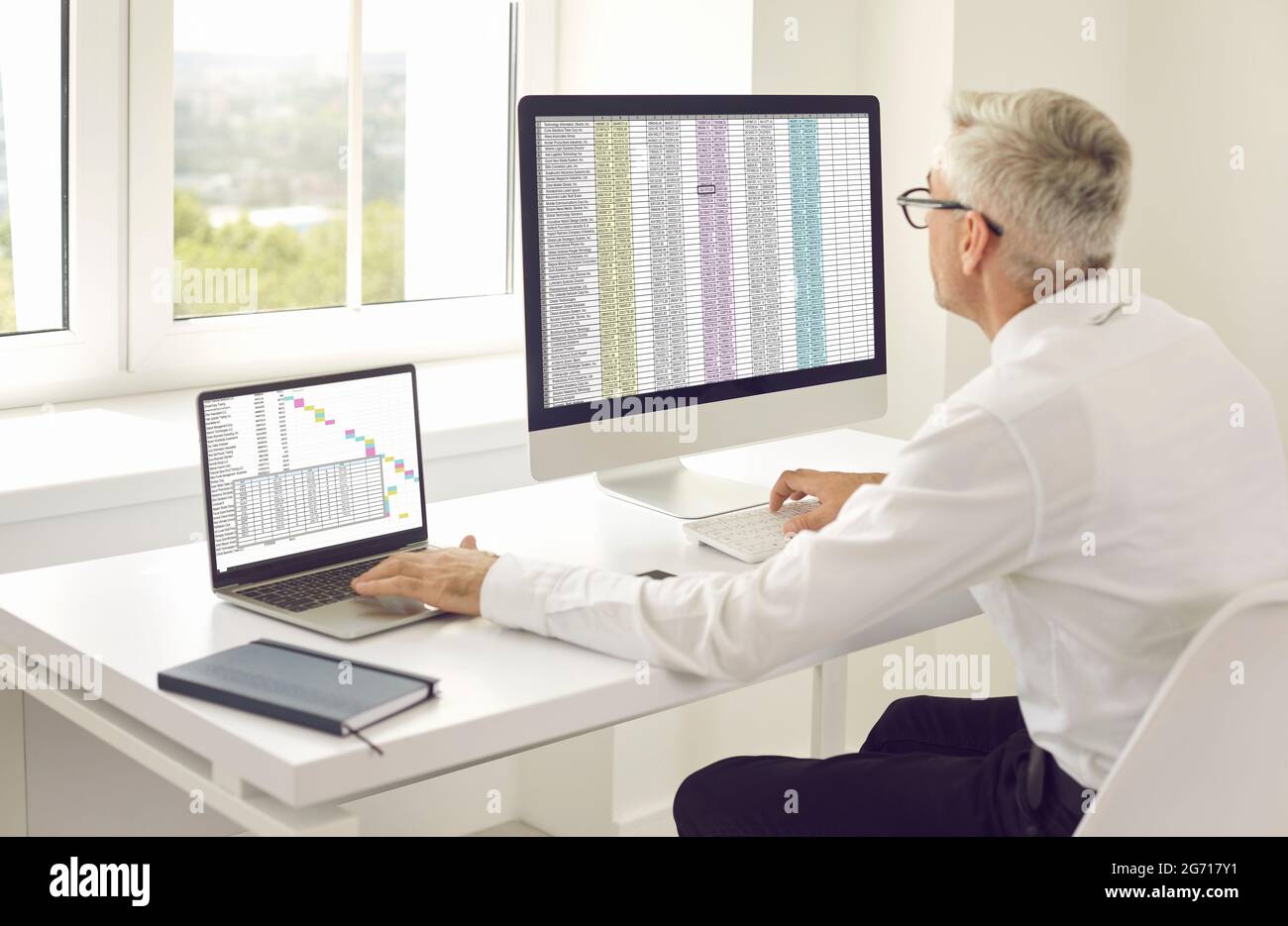 Uomo seduto alla scrivania dell'ufficio e che fa una cartografia Gantt utilizzando un computer portatile e un computer desktop Foto Stock