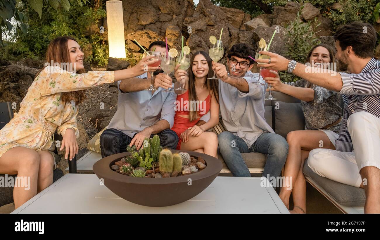 Gruppo di giovani che si divertono a raccogliere cocktail e a tostare insieme seduti su un divano in giardino. Cocktail party. Mettere a fuoco sugli occhiali. Foto Stock