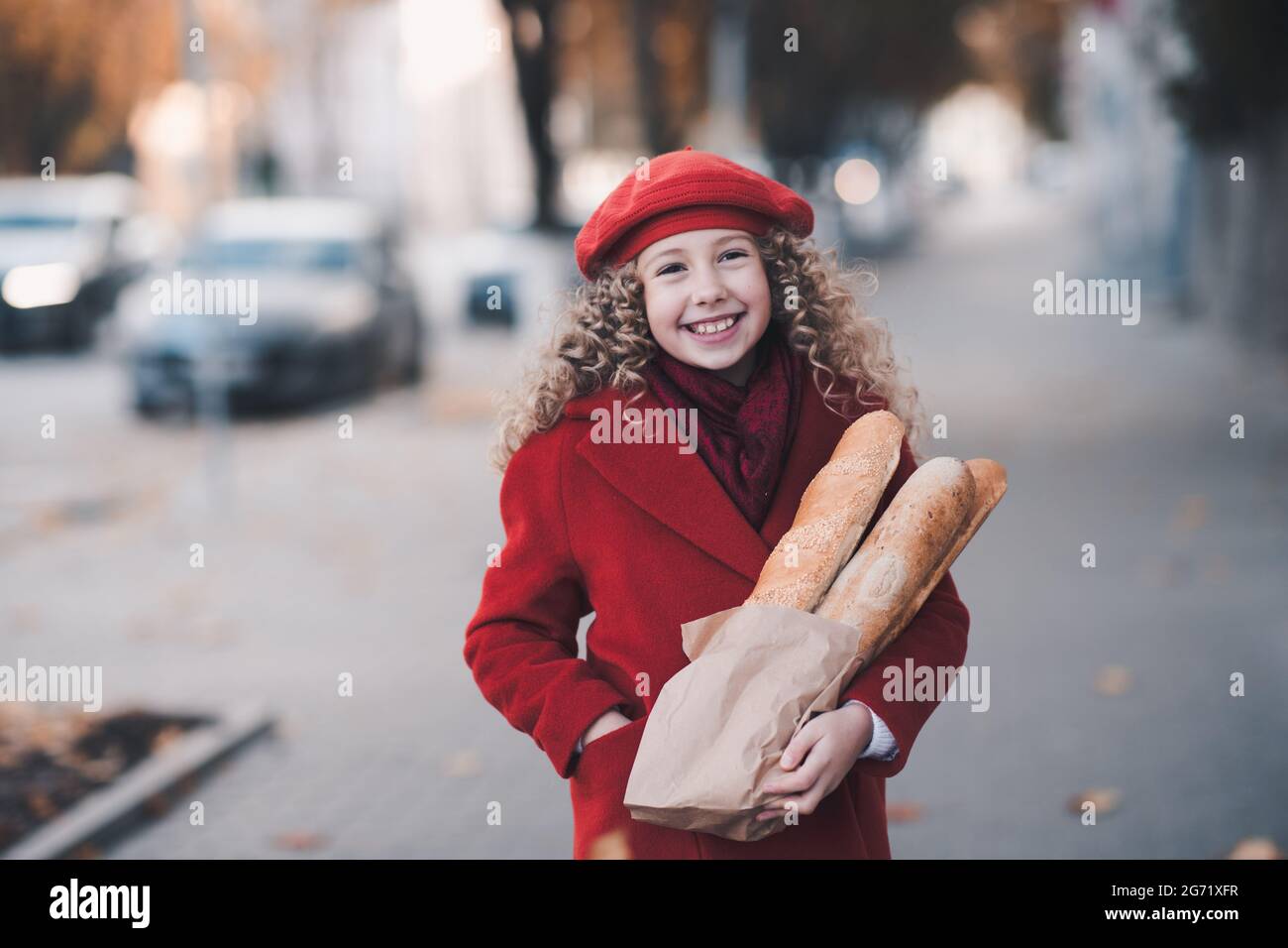 Sorridente ragazzina 5-6 anni tenere pasta di pane fresco indossare cappotto rosso elegante in strada all'aperto. Stagione autunnale. Infanzia. Messa a fuoco selettiva. Foto Stock