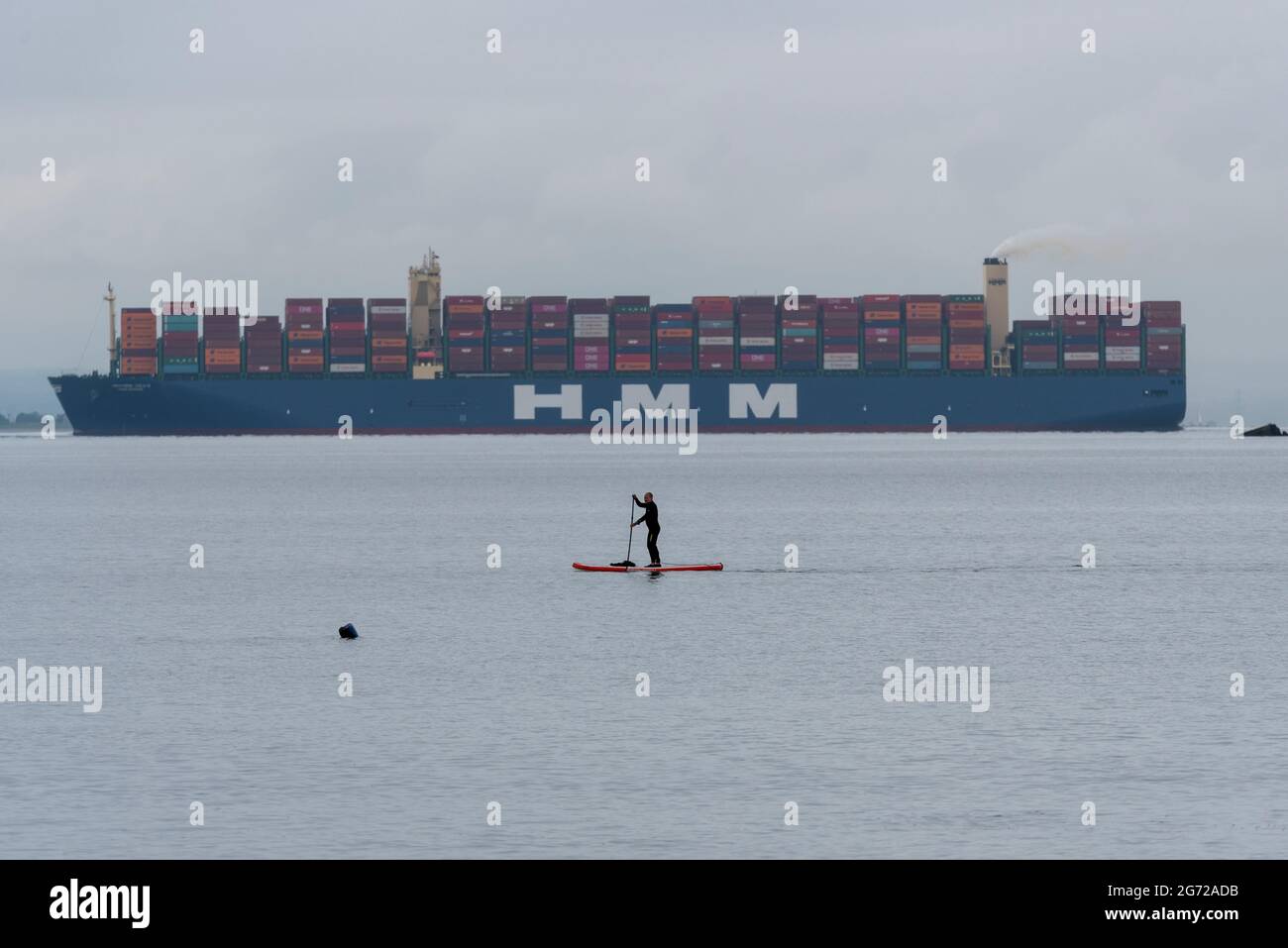 Shoeburyness, Essex, Regno Unito. 10 luglio 2021. Una persona pagaia su una tavola a pale in piedi sull'estuario del Tamigi mentre l'HMM Gdansk passa sulla sua strada verso il Mare del Nord. Traffico misto di fiume Foto Stock