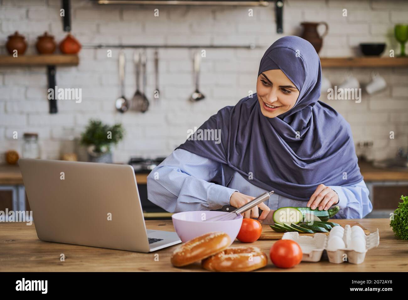 Casalinga musulmana che cucina un pasto e guardare un film su un computer portatile Foto Stock