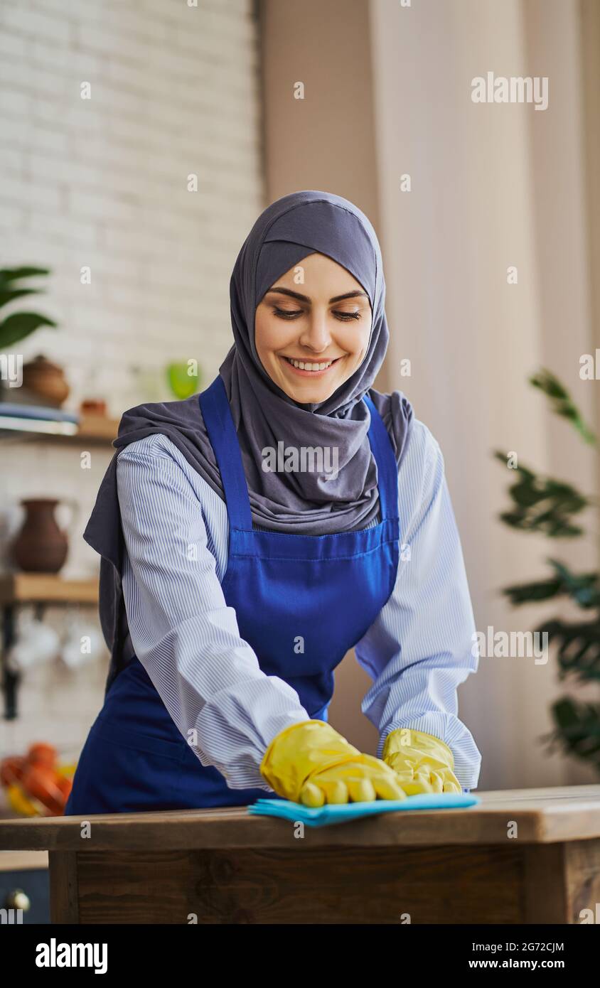 Bella donna araba in grembiule e guanti per la pulizia di un tavolo in cucina Foto Stock