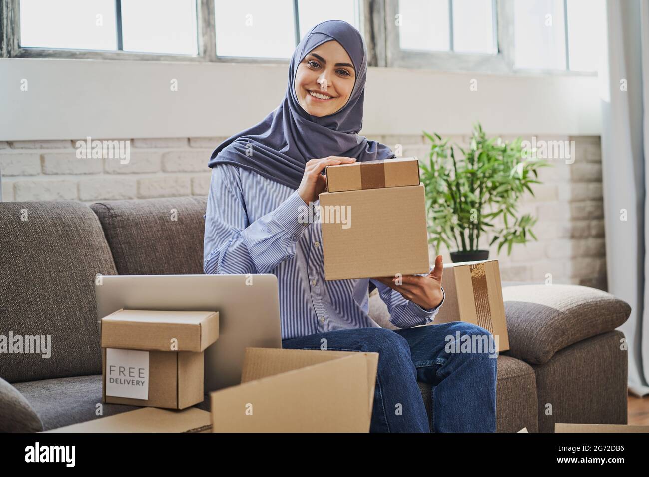 Donna musulmana seduta sul divano con un computer portatile e mostrando scatole di cartone Foto Stock