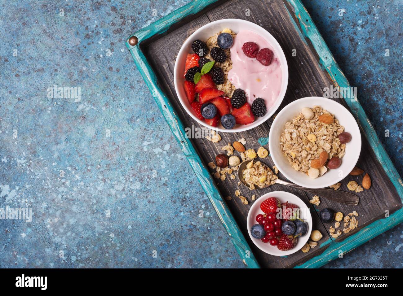 Vista dall'alto della ciotola con scaglie di avena, lampone, fragola, mirtillo, ribes rosso, yogurt fatto in casa alla mandorla, alla nocciola e ai frutti su vassoio turchese e blu Foto Stock