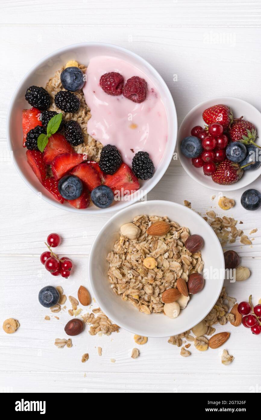 Vista dall'alto di ciotole con scaglie di avena, lampone, fragola, mirtillo, ribes rosso, mandorla, nocciola e yogurt rosa di frutta fatti in casa su sfondo di legno bianco Foto Stock