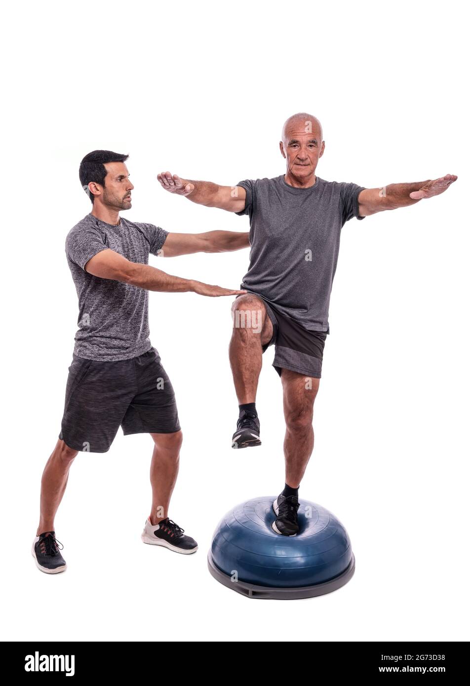 Un uomo anziano che fa un esercizio di equilibrio, su una gamba, su una palla emisferica. Con l'aiuto di un fitness trainer. Su uno sfondo bianco isolato. Foto Stock