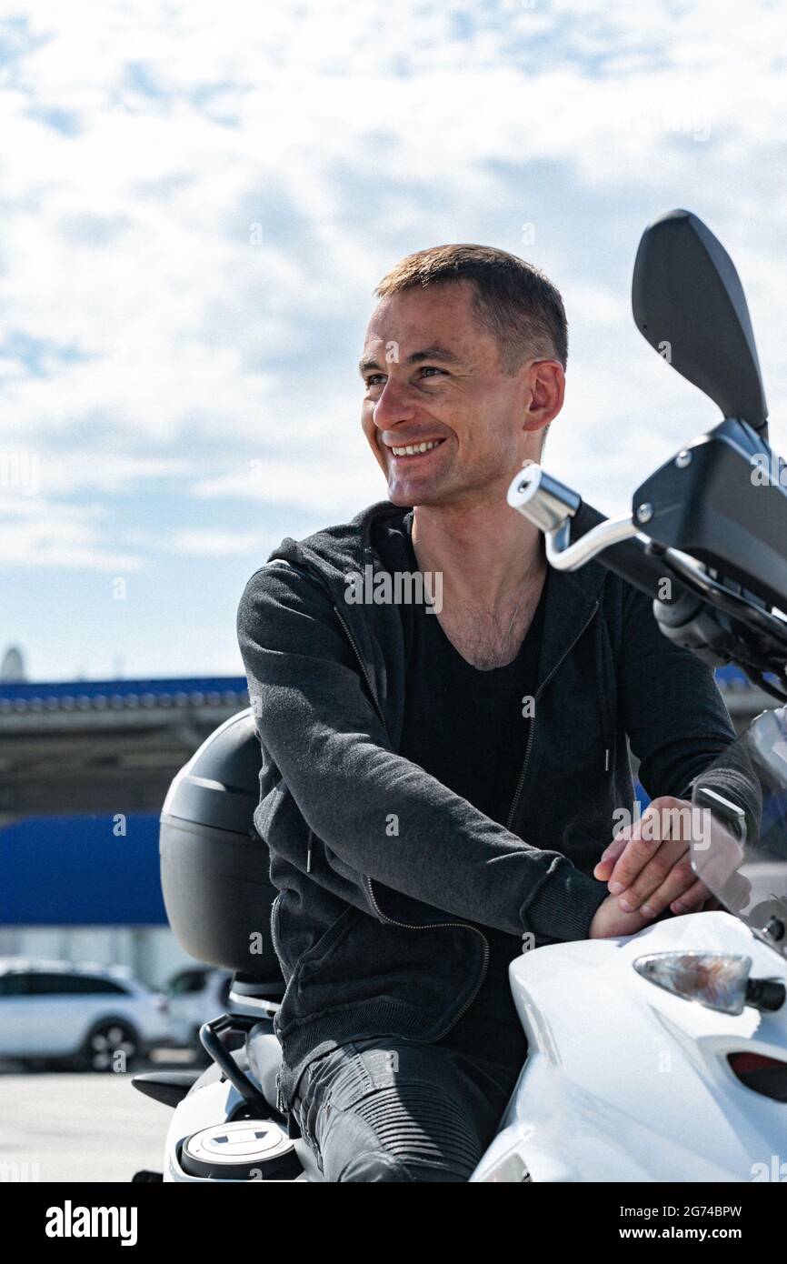 Giovane ed elegante uomo sorridente seduto su una motocicletta. Ritratto di un bel biker che si posa su una bicicletta in una giacca nera in pelle. Stile di vita phot Foto Stock