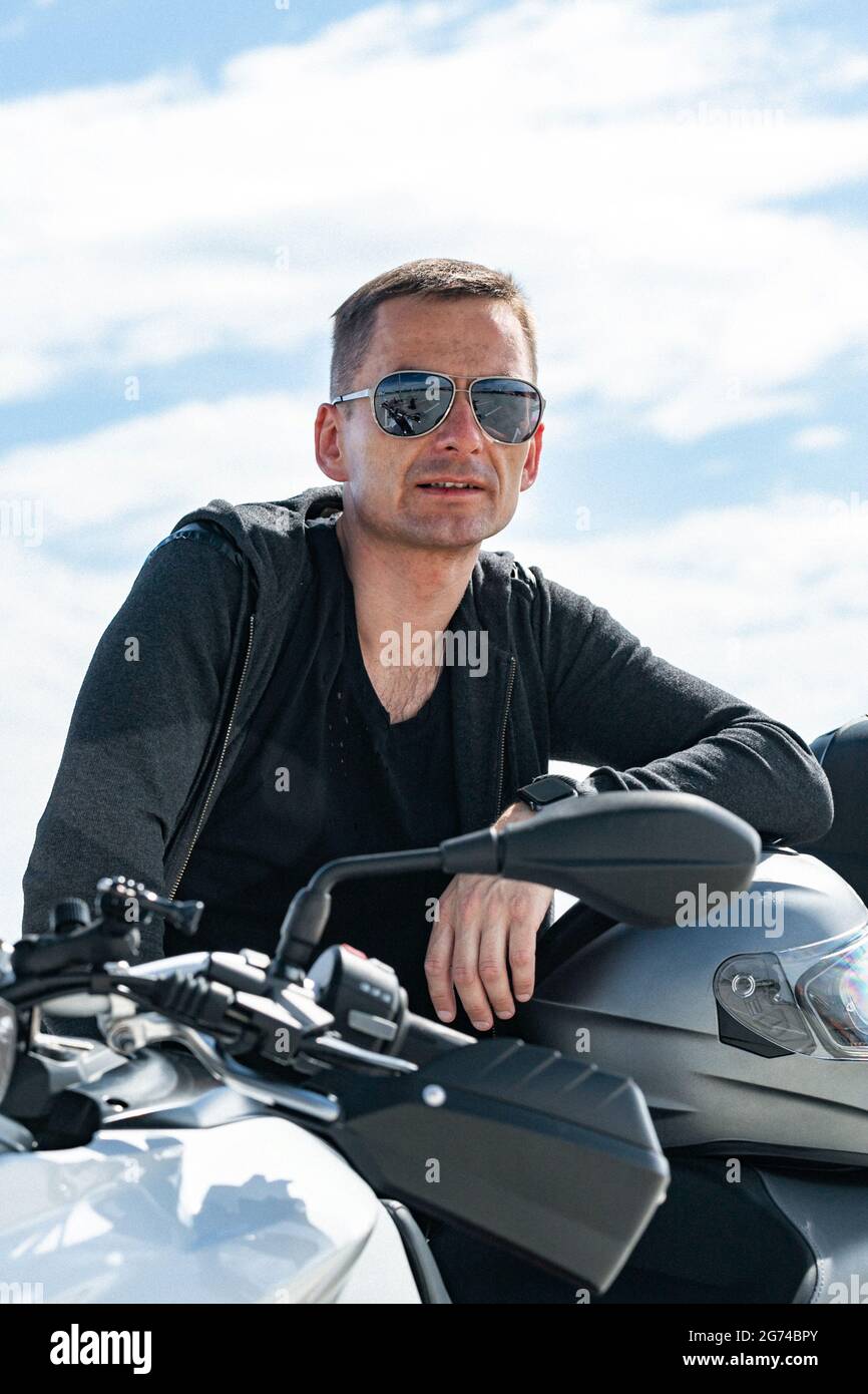 Attraente giovane uomo di stile in occhiali da sole si siede su una motocicletta. Ritratto bello biker in posa su una bicicletta in una giacca nera in pelle. Foto stile di vita Foto Stock