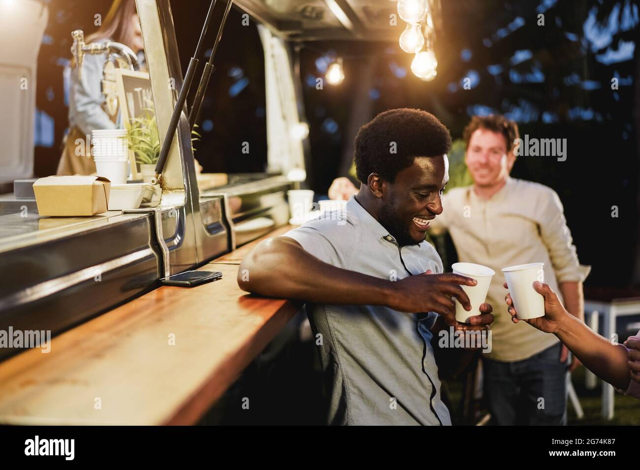 Persone multirazziali che si acclamano con le bevande al bancone al ristorante di camion cibo all'aperto - Focus sulla faccia dell'uomo afroamericano Foto Stock