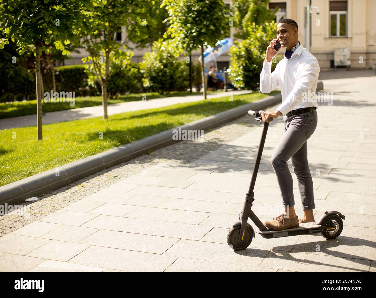 Bel giovane afroamericano che usa il telefono cellulare mentre si è in piedi con uno scooter elettrico su una strada Foto Stock