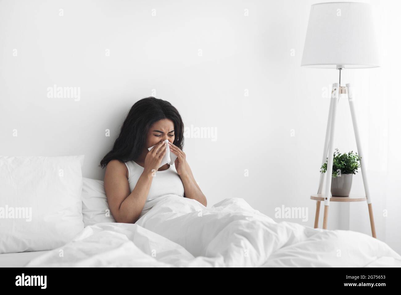 Giorno di malattia a casa. Malata donna afro-americana ha naso che cola, tosse e freddo, naso che soffia, seduta a letto, spazio copia Foto Stock