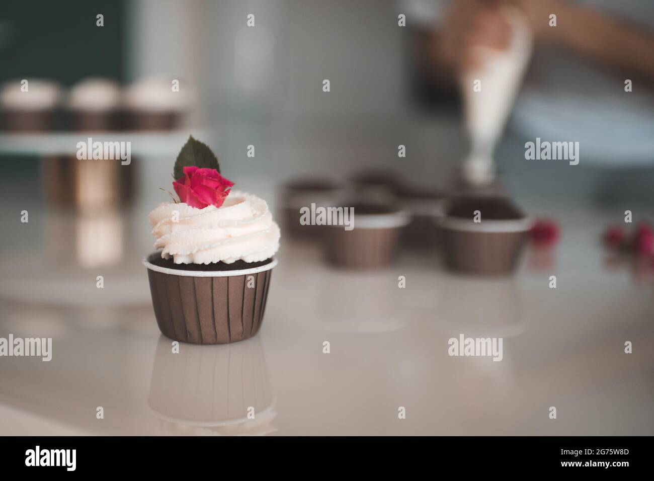 Preparare deliziosi cupcakes di cioccolato con panna montata e fiori freschi decorazione rosa soggiorno sul tavolo da cucina primo piano. Preparatevi per la festa di compleanno. Vai Foto Stock