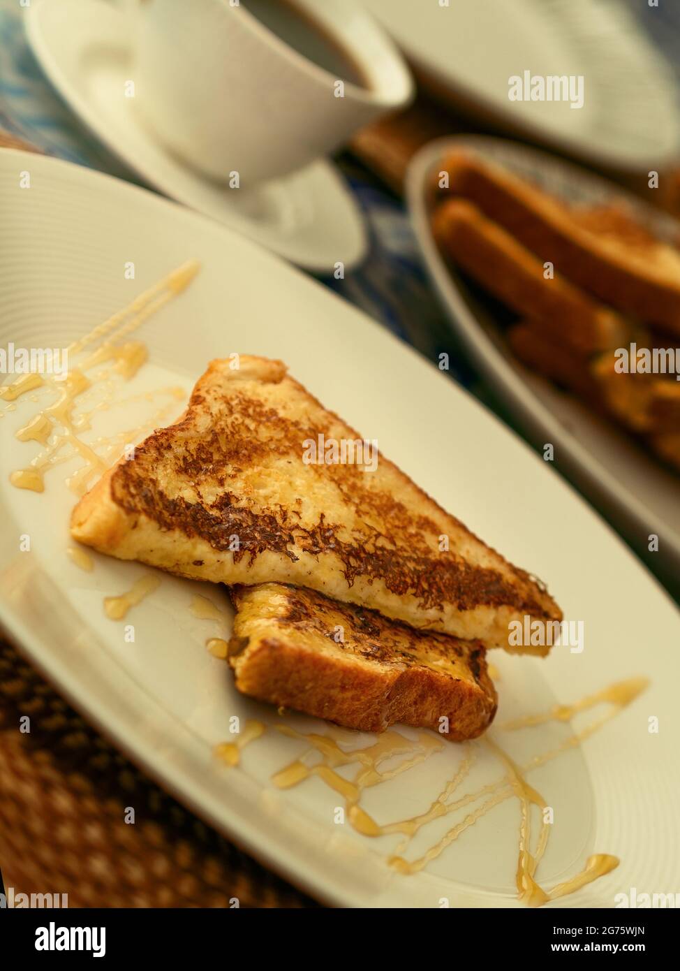 Due toast alla francese serviti con miele su un piatto bianco per colazione o brunch, sullo sfondo una tazza di caffè nero e una pila di toast alla francese. Foto Stock