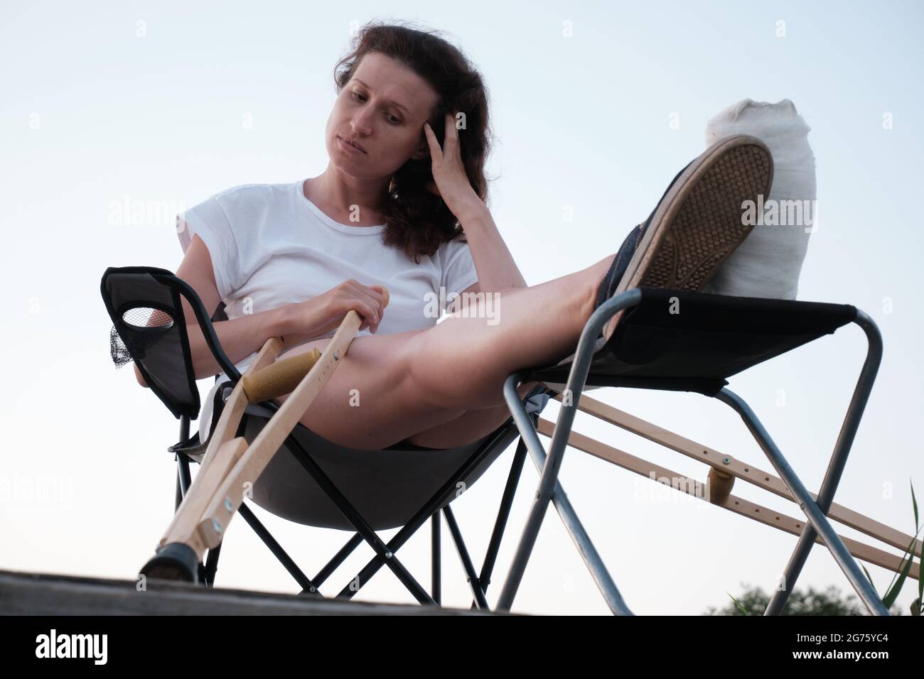 Donna con rotto in cast è seduta su sedia in natura. Lesioni alle gambe. Perdita di mobilità. Riabilitazione dopo una frattura. Foto Stock