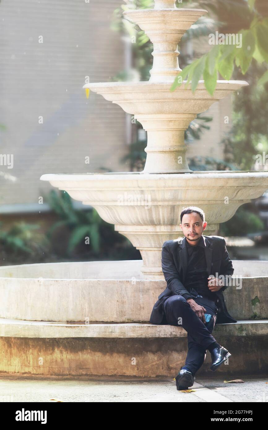 Attraente uomo d'affari in un suit.Sit nero nel sole di sera nel mezzo della natura e trees.There è una grande fontana sullo sfondo. Foto Stock