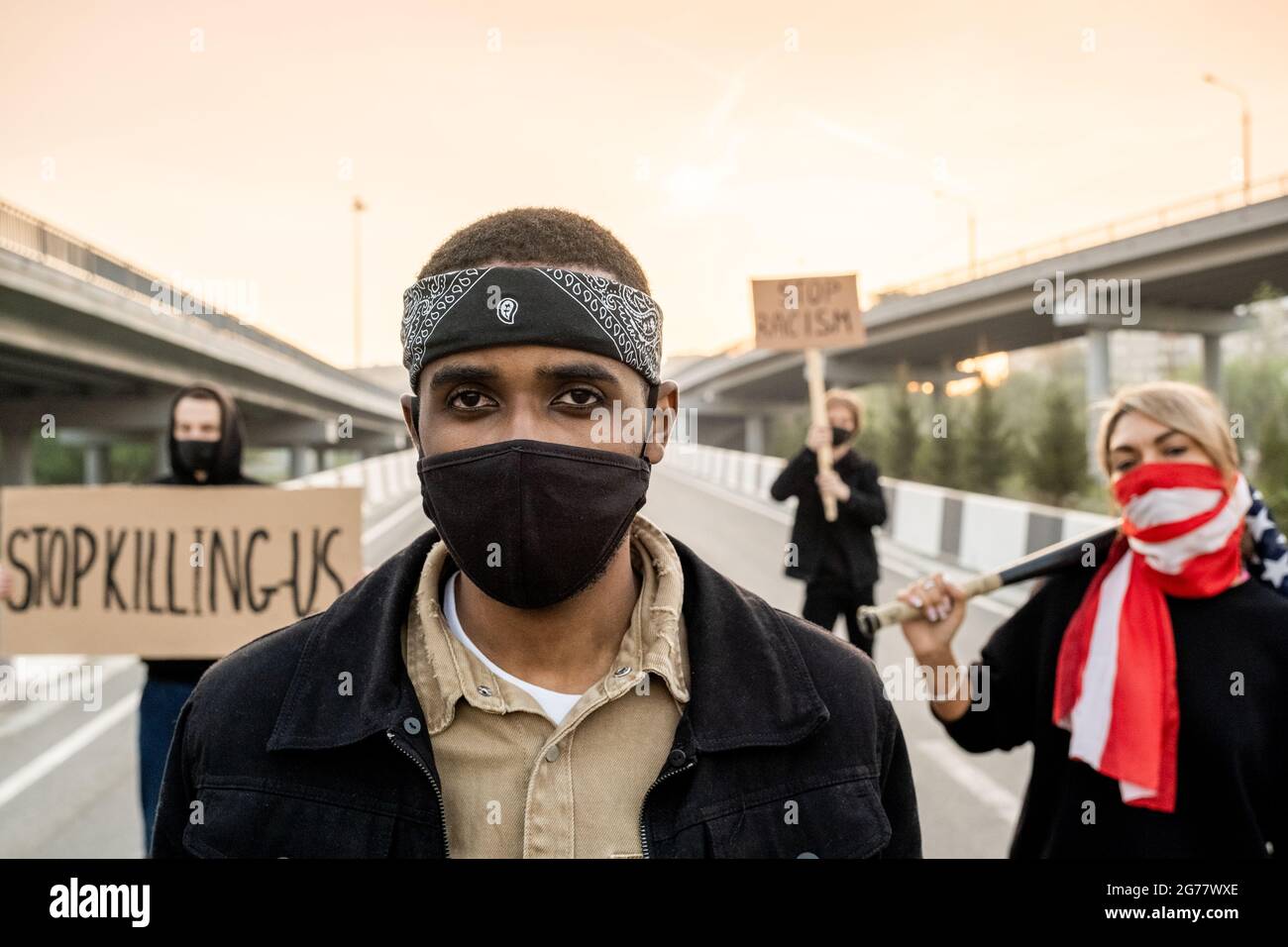 Ritratto di un uomo brutale serio in maschera con archetto e maschera facciale in piedi per strada contro la protesta di persone con segni Foto Stock