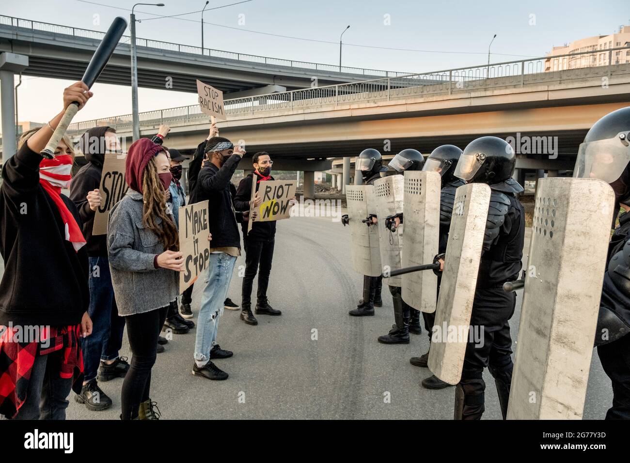 Gruppo di giovani manifestanti maschere che tengono i segni e si levano in piedi davanti alle guardie della polizia con gli scudi Foto Stock