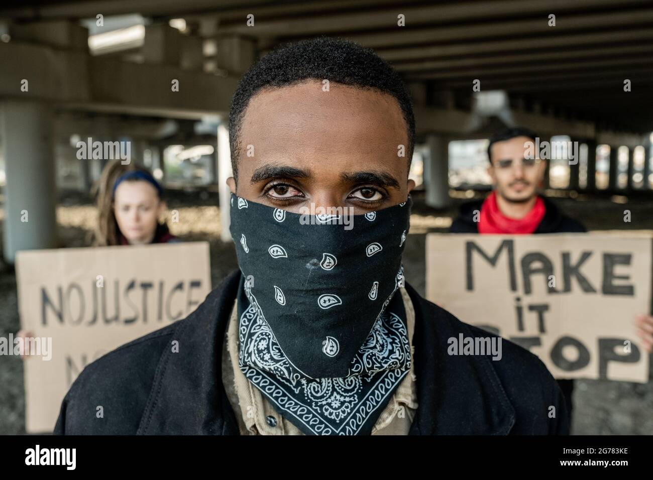 Ritratto del giovane protestore nero in bandana sul volto in piedi contro persone con segni sotto il ponte Foto Stock