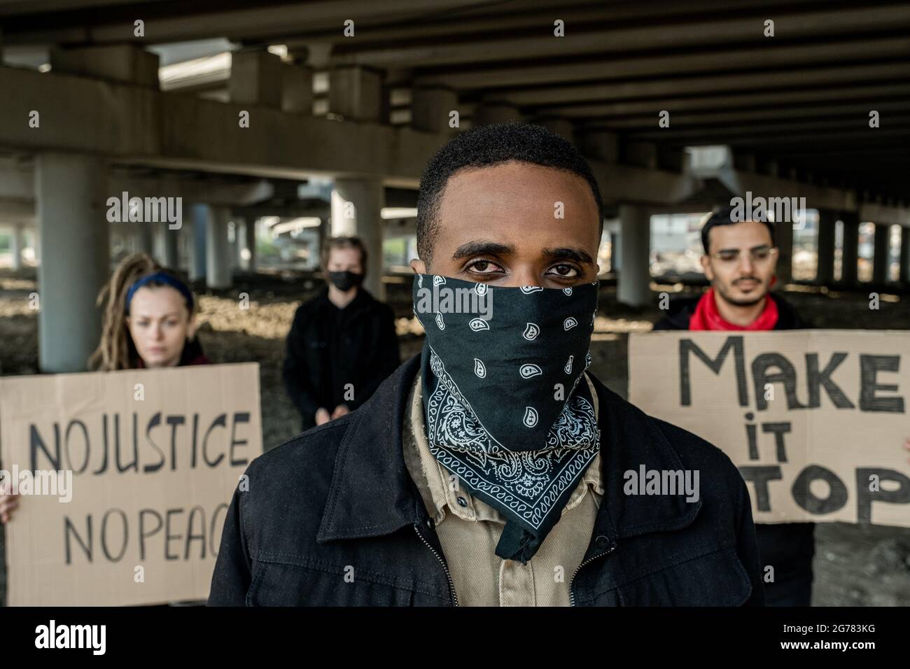 Giovane uomo nero in bandana in piedi contro la protesta di persone con striscioni sotto ponte Foto Stock