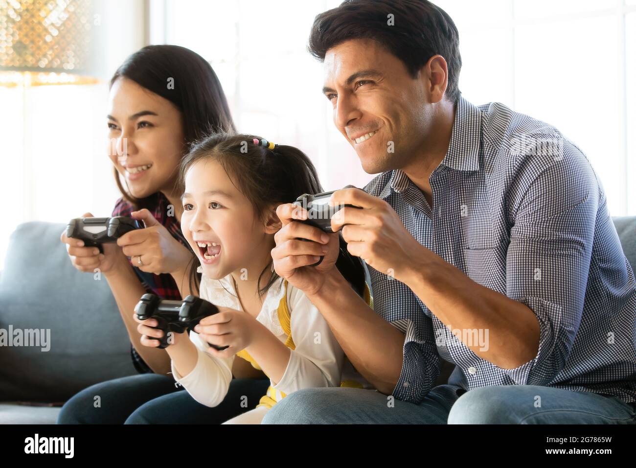 Diversità multirazziale caucasico e asiatico famiglia padre, madre e figlia seduti insieme sul divano e giocare gioco al computer insieme con divertimento e. Foto Stock