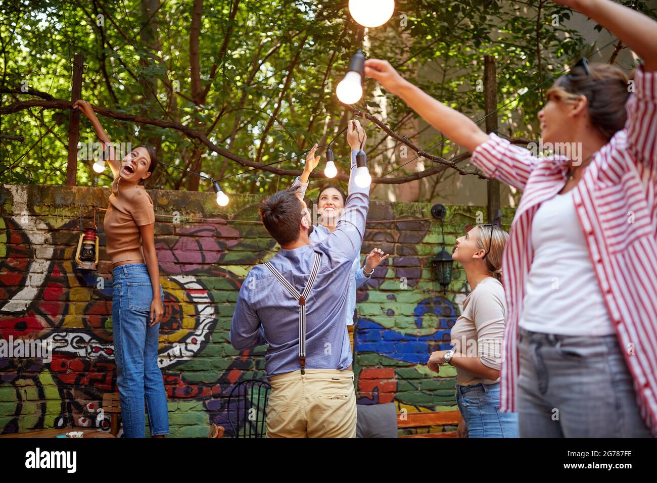 gruppo di giovani adulti caucasici uomo e donne che allestiscono lampade appese che si allungano, ridono, si preparano per la festa Foto Stock