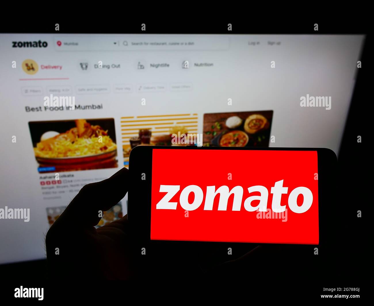 Persona che tiene il cellulare con il logo della società indiana di ordinazione alimentare online Zomato sullo schermo di fronte alla pagina web business. Mettere a fuoco il display del telefono. Foto Stock