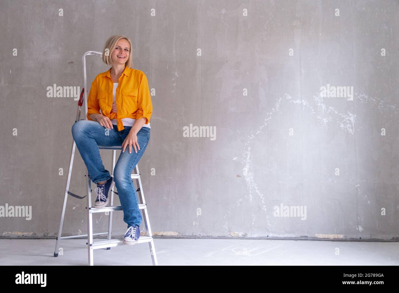 Appartamento di ristrutturazione. Donna felice si siede sulle scale vicino a muro vuoto a casa. Muro libero, ripara casa con le proprie mani. CopySpace per il testo. Foto Stock