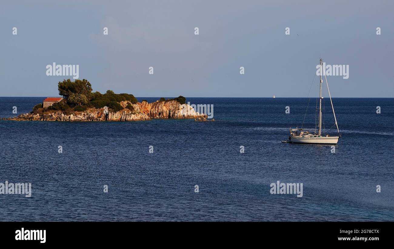 Isole IONIE, Ithaca, isola di Odysseus, costa nord-orientale, Baia di Marmakas, Isolotto di Agios Nikolaos dalla spiaggia, mare blu scuro, cielo azzurro chiaro, cappella con tetto rosso sulla sinistra dell'isola, alberi e altre vegetazione accanto ad essa, una piccola barca a vela ancorano alla destra dell'isolotto Foto Stock